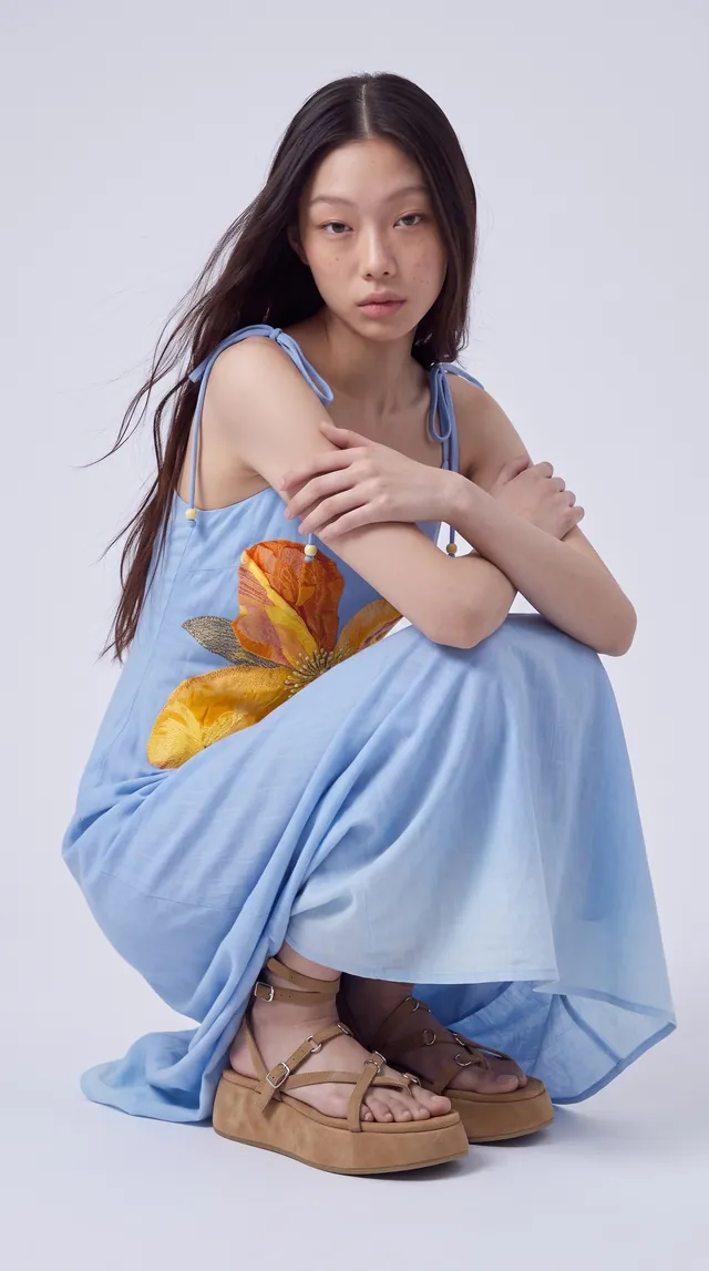 AI-generated still: the same model seated, showing the dress print and sandals